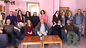 Group of people, including nuns and laypersons, posing for a photo in a pink-walled living room with a wooden coffee table and bookshelf in the background.