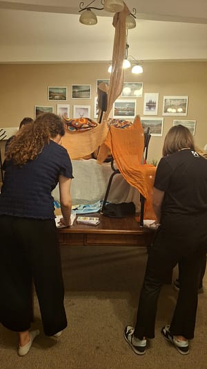 People assemble a fabric-covered sculpture in a living room-like space, orange cloth draped over chairs and a tall central form amid artwork on the wall behind them.