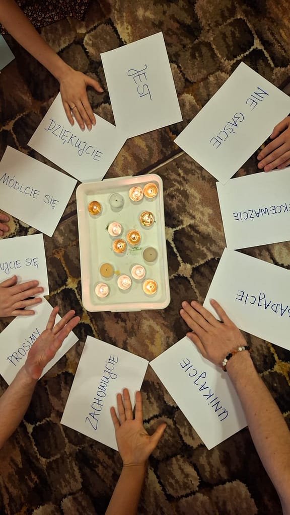 People sit in a circle on a patterned rug, a tray of lit tealight candles in the center, with white cards bearing handwritten Polish phrases around them.