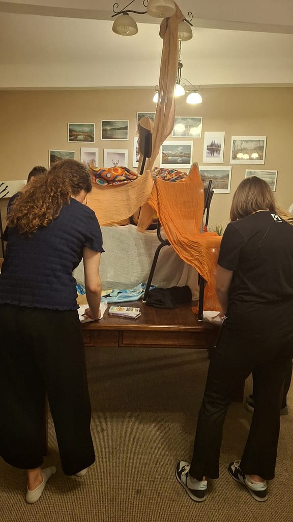 People assemble a fabric-covered sculpture in a living room-like space, orange cloth draped over chairs and a tall central form amid artwork on the wall behind them.