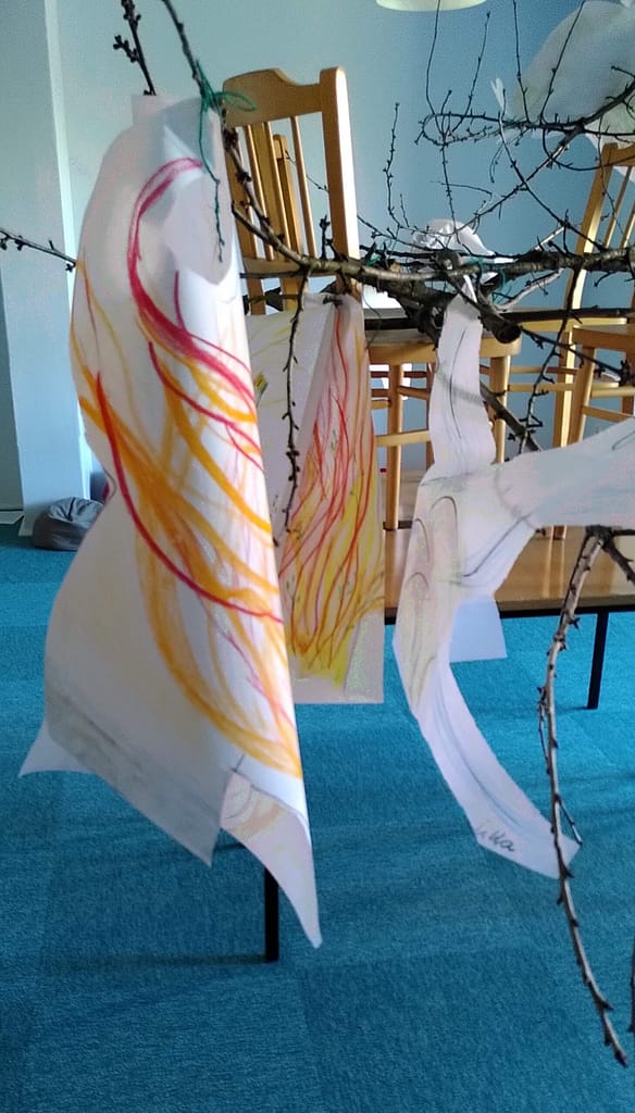 Art installation with colorful drawn fabric strips hanging from branches in a bright blue room.