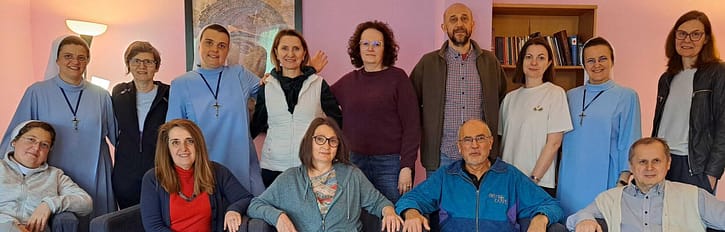Group of fourteen adults posing in a pink-walled room, some seated on a blue couch and others standing, smiling at the camera.