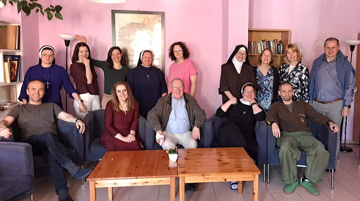 Group of people, including nuns and laypersons, posing for a photo in a pink-walled living room with a wooden coffee table and bookshelf in the background.