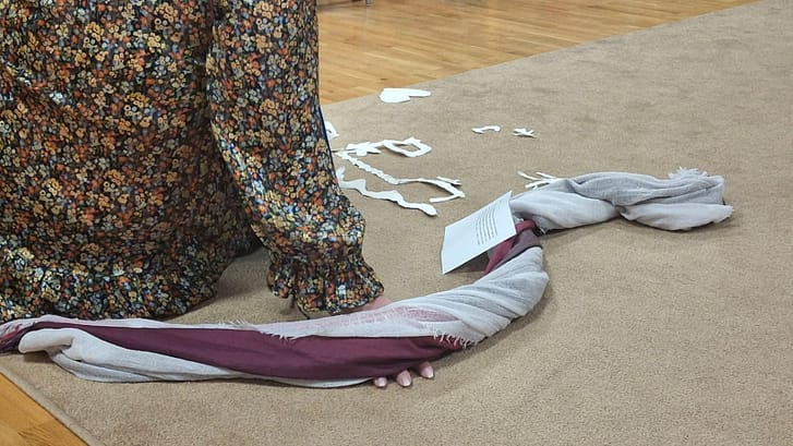 Person in a long colorful floral dress kneels on a beige carpet, with a grey and maroon scarf laid out in a loop and small torn white paper shapes scattered nearby.
