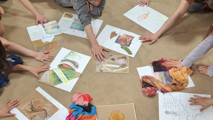 Group of people sitting on a carpet in a circle, each with colorful drawings and papers laid out between them, hands reaching toward the center.