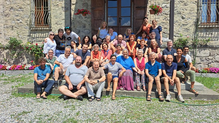 Large group of people posing on stone steps in front of a rustic stone house with wooden doors and hanging flower baskets, smiling at the camera.